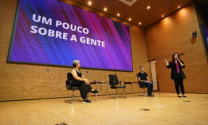 Foto da Elza Maria Albuquerque em pé no palco do auditório do Tribunal de Justiça Federal de Tocantins. Atrás dela, há duas pessoas sentadas: Adriana Magna Ramalho e Leonardo Gleison. Ao fundo, há um telão com texto.